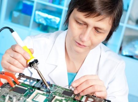 A person in a lab coat uses a soldering iron on a circuit board, focusing intently on the work.