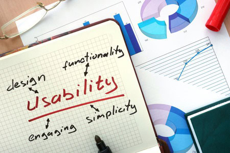 Notebook with "usability" and related terms like design, functionality, engaging, simplicity. Background shows graphs and charts. Glasses and pen are placed nearby.