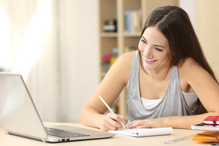 A woman sits at a desk, smiling while writing in a notebook, with an open laptop in front of her.