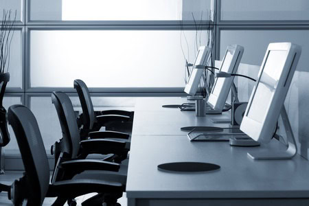 Empty office with four ergonomic chairs at desks, each with a computer monitor. The background features frosted glass panels.