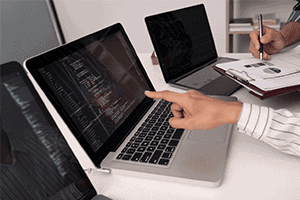 Close-up of a person pointing at a laptop screen displaying code, surrounded by other laptops and a clipboard with documents on a desk.