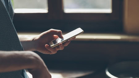 Person holding a smartphone, standing near a window with sunlight streaming in.