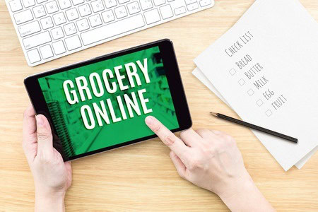 Person holding a tablet displaying "Grocery Online" next to a checklist on paper with items like bread and milk, and a keyboard in the background.