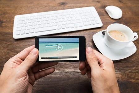 Person holding a smartphone with a beach video paused on the screen. A keyboard, mouse, and cup of coffee are on a wooden table.
