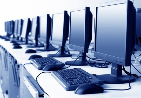 A row of desktop computers with keyboards and mice on a long table, arranged in an office or computer lab setting.