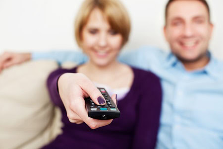 A person holds a TV remote control while sitting on a couch next to another person. Both are smiling.